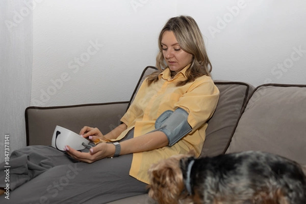 Obraz A woman sits on a couch and uses a blood pressure monitor. A dog is on the floor nearby. The setting looks casual and homey during daylight hours.