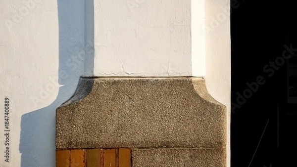 Fototapeta Minimalist architectural close-up of a white plastered wall meeting a textured stone pillar base. Natural sunlight creates sharp geometric shadows on the clean building exterior.