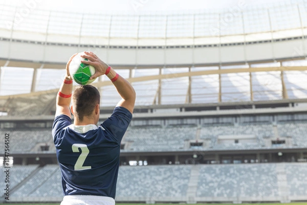 Obraz Athlete is standing on grass field holding green and white ball overhead, wearing navy jersey 2