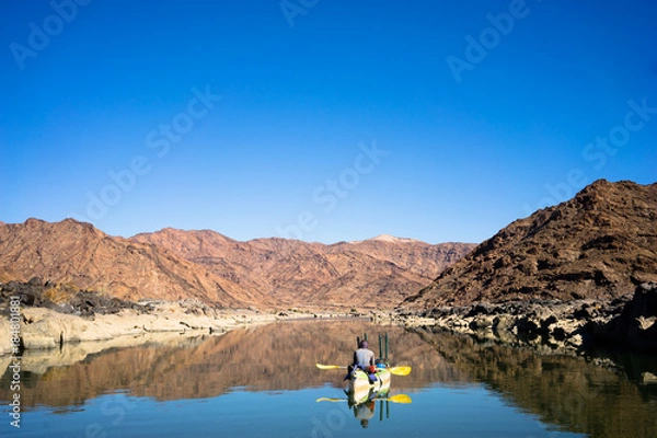 Fototapeta A canoe drifting along the calm waters of the Orange river with rugged mountains reflecting off the calm water
