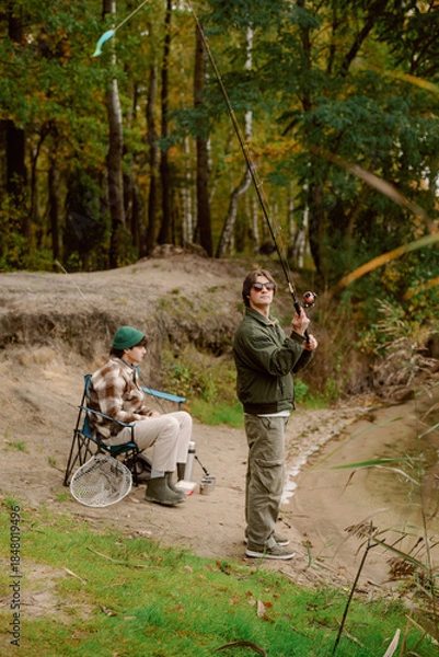 Obraz Two friends enjoy a peaceful afternoon fishing by a quiet river in the forest. One is casting a line while the other relaxes, surrounded by vibrant trees and nature.