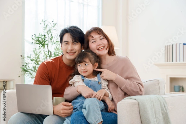 Fototapeta Happy Asian family of three, a father, mother, and young daughter, smiling and sitting on a sofa with a laptop at home, enjoying a moment of family bonding and connection.