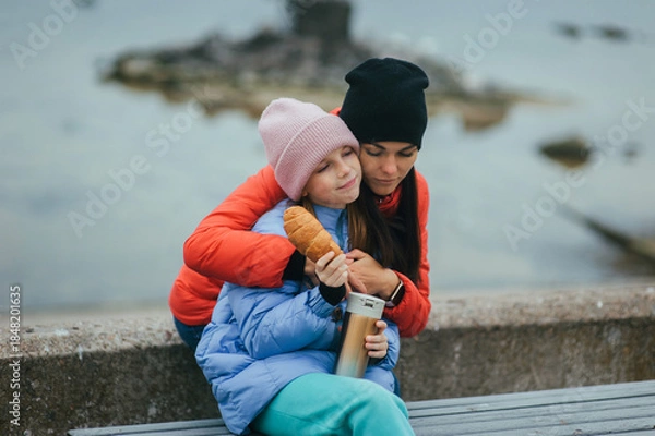 Obraz Photo, portrait of a beautiful teenage girl, child with a thermos of hot tea and croissant, food, sitting with her mother, young woman on a bench with the sea in the background on a picnic.