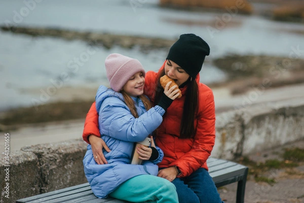 Obraz Photo, portrait of teenage girl, child with thermos of hot tea and croissant, food, sitting with mother, young woman on bench against sea background on picnic in autumn.