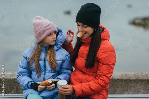 Obraz Photo, portrait of teenage girl, child with thermos of hot tea and croissant, food, sitting with mother, young woman on bench against sea background on picnic in autumn.