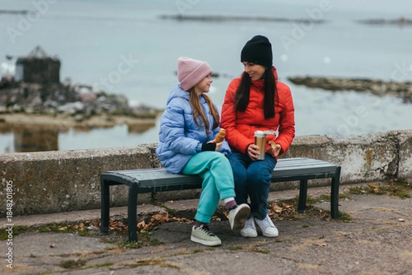 Obraz Photo, portrait of a beautiful teenage girl, child with a thermos of hot tea and croissant, food, sitting with her mother, young woman on a bench with the sea in the background on a picnic.