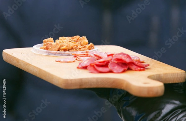 Obraz Tasting samples of sliced sausage and traditional pork cracklings served on a wooden board at the Naplavka farmers market in Prague, inviting visitors to try local cured meats.