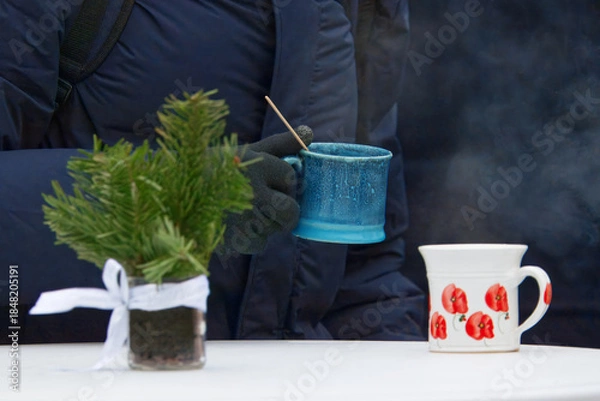 Obraz Cold winter morning at Prague Naplavka farmers market. A bundled visitor enjoys a steaming hot drink at a simple table, advent greenery adding warmth, comfort and seasonal charm to the scene.