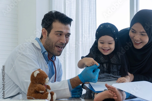 Obraz Friendly male doctor playing with cute Muslim little girl patient and mother in clinic. Happy healthcare professional interacting with kid near teddy bear for pediatric concept.