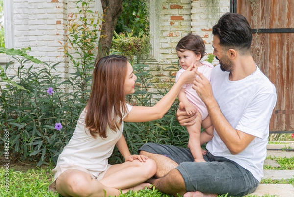 Obraz Happy multi-ethnic family sitting on green grass in garden. Middle Eastern father and Asian mother playing with cute baby daughter. Mixed race parents lifestyle concept with love.