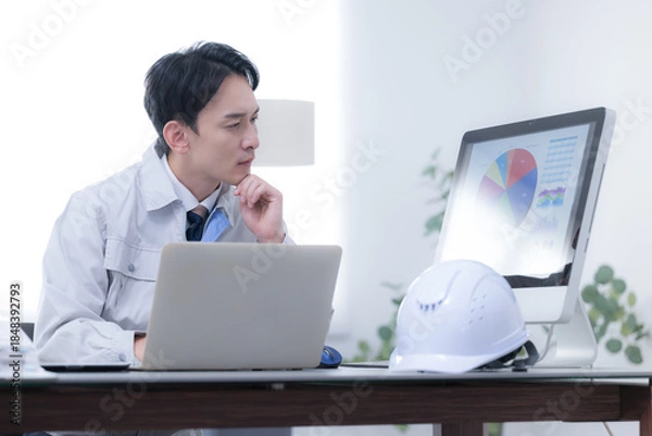 Obraz 現場事務所で考える作業着の作業員　発注や下請けで納期に困るや経営などの戦略を考える