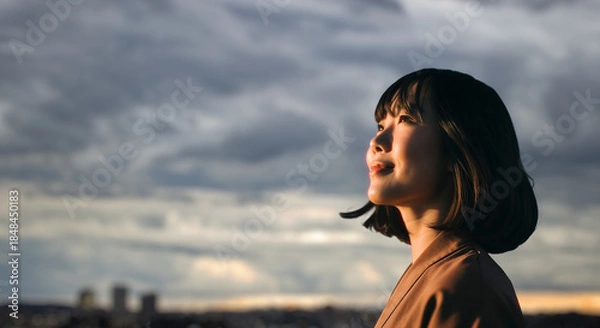 Obraz 美しい夕焼け・朝焼け　夕日・朝日に照らされた空と街が日の出の光を浴びた背景　空を見上げ上を向きリラックスした表情の女性の横顔　日本・ビジネス・転職出勤・旅行・観光・アウトドア・リラクゼーションの背景　希望・安堵のイメージ
