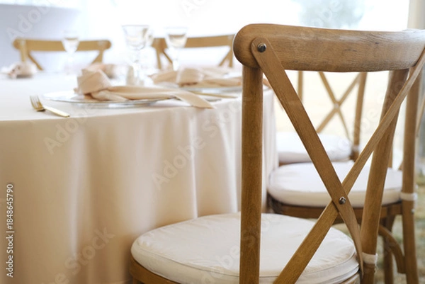 Fototapeta Elegant wooden chairs arranged around a beautifully set wedding banquet table with soft linens and glassware, creating an inviting atmosphere for celebration and joy