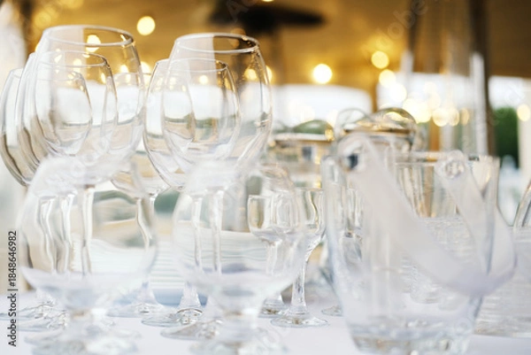 Fototapeta Glassware collection including wine glasses, tumblers, and a pitcher arranged on a table with blurred background lights and soft bokeh effect during an outdoor event