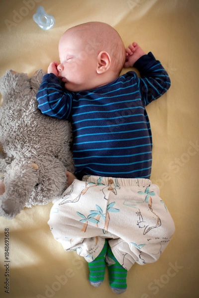 Fototapeta Cute infant sleeps peacefully on a yellow blanket, wearing striped shirt and patterned pants. The little one rests next to a gray stuffed animal and a pacifier.