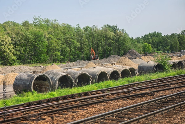Obraz Concrete pipes and gravel piles line a construction site beside active railway tracks, excavator working near forested backdrop. Scene blends transportation infrastructure with utility development