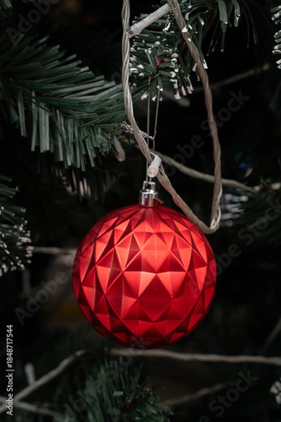 Fototapeta Festive Bauble: A radiant red ornament, adorned with a geometric pattern, glistens gracefully amidst the lush, dark green foliage of a Christmas tree.