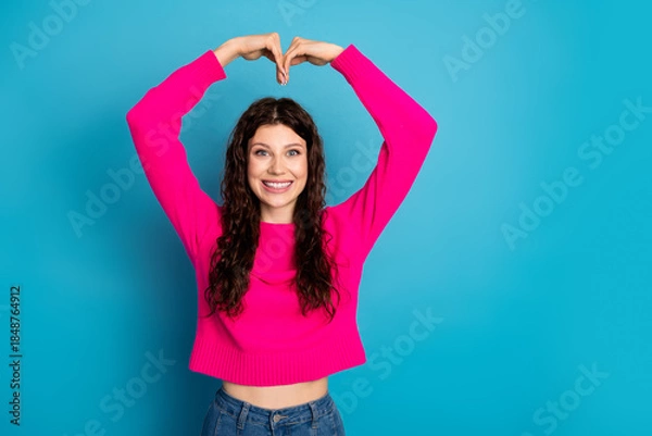 Fototapeta Young woman in pink sweater forms a heart with her arms against a blue background