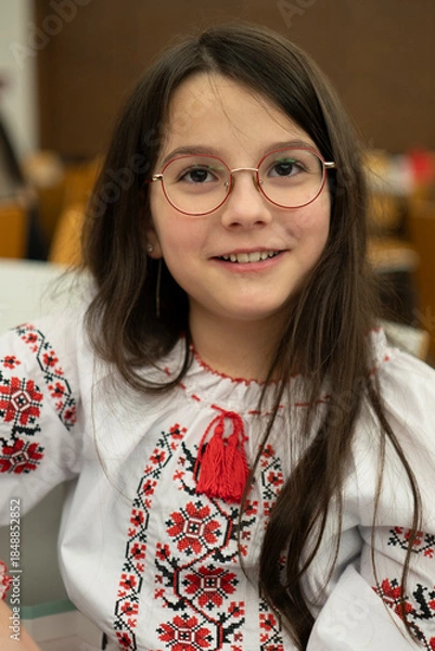 Fototapeta Vertical photo of smiling schoolgirl in glasses wearing vyshyvanka sitting in classroom, education concept