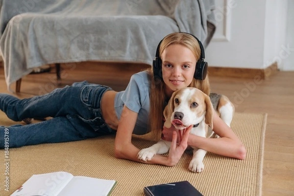 Obraz Looking forward, in headphones, embracing animal. Happy girl is on the floor at home with dog, domestic room