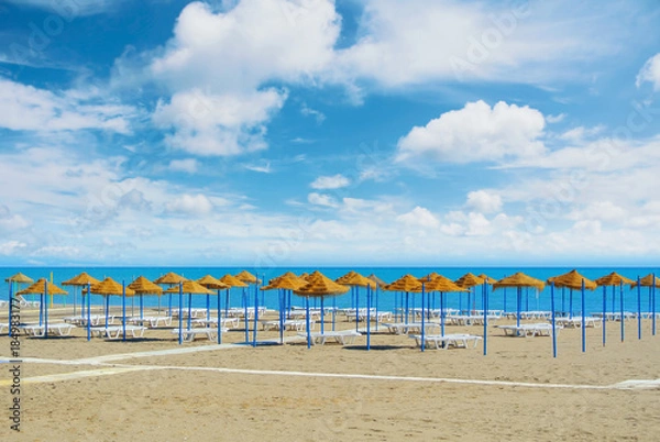 Fototapeta Empty perfect dreamy paradise beach with umbrellas tents made of palm leaves and white beach lounge chairs standing at the sandy bay by the blue sea and beautiful cloudy sky. Vacation holidays concept