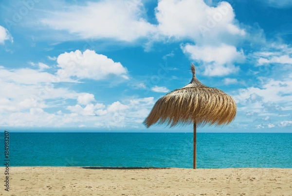 Fototapeta Beach umbrella made of palm leaves standing at the perfect dreamy paradise white yellow sandy bay beach by the blue sea and beautiful dramatic cloudy sky. A place for relax and vacations by the water.