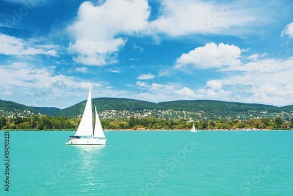 Fototapeta Panoramic view of lake river blue transparent water, a white sport modern luxury yacht sail boat floating and a green shore with forest, hills, villages and beach. Holiday by the water and sail race.