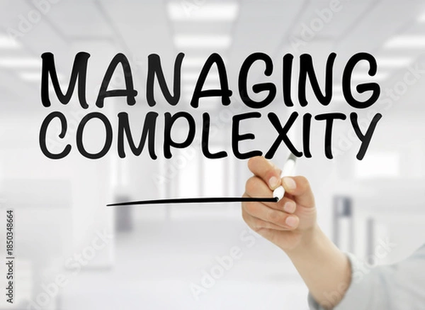 Obraz A conceptual business image showing the phrase managing complexity handwritten in bold black letters on transparent glass, with a blurred modern white office interior in the background. The clean corp