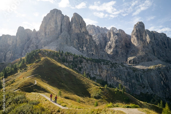 Fototapeta Two hikers walking into the mountains along a small trail with a beautiful panorama of high rocky mountain peaks and green fields and trees ahead of them