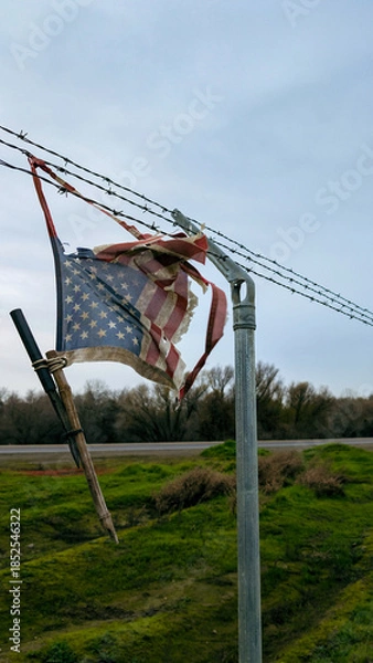Obraz A tattered American flag remains snagged on a weathered barbed-wire fence