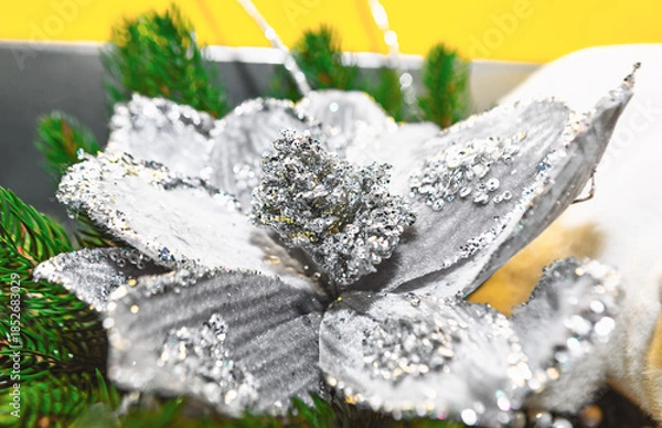 Obraz Glittering silver artificial flower with sequined petals rests among green pine branches, evoking Christmas festive charm. Bright and soft white tones in background enhance its decorative appeal