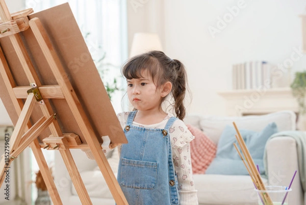 Fototapeta Serious little girl concentrating on painting at wooden easel in living room creative hobby and child development concept