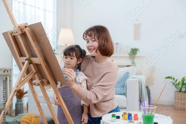 Fototapeta A happy Asian mother and her young daughter smiling while painting on an easel at home, enjoying a creative art activity and family time.