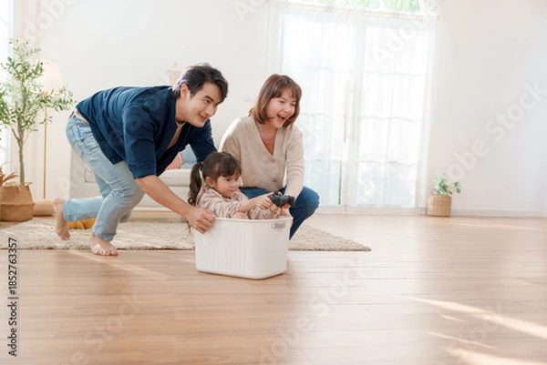 Fototapeta Happy Asian parents pushing their little daughter in a plastic basket while she holds a game controller at home Fun family bonding and creative childhood play in a bright modern living room
