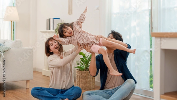 Fototapeta A delighted Asian mother and father lifting their cheerful young daughter high in the air, simulating flight during playtime in their brightly lit living room, embodying family happiness.