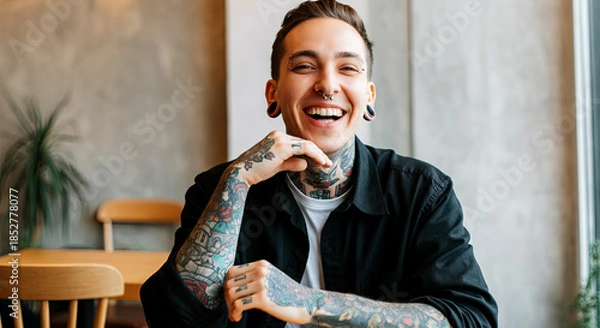 Fototapeta Smiling young man with tattoos, wearing a black shirt, sitting at a wooden table in a cozy cafe, showcasing a joyful expression and relaxed atmosphere