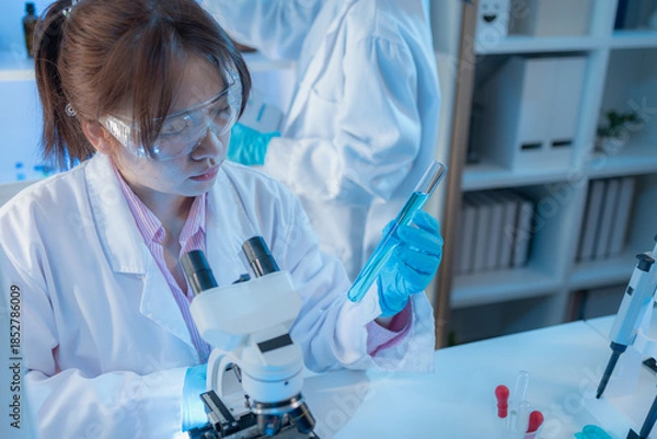 Obraz Two female scientists in white lab coats working together in a bright laboratory with microscopes, glassware, and blue test tubes, representing teamwork and modern scientific research environment.