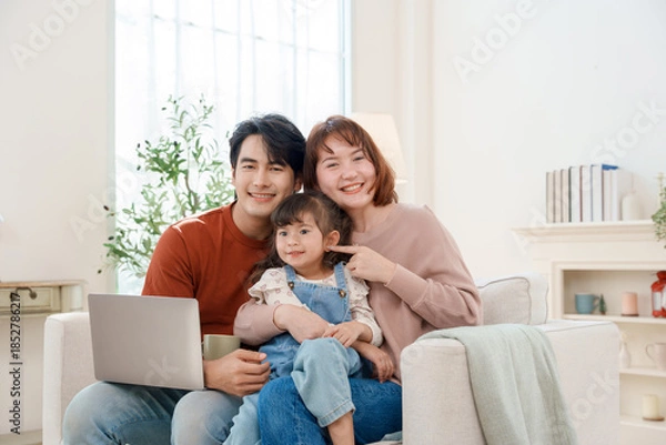 Fototapeta Happy Asian family of three, a father, mother, and young daughter, smiling and sitting together on a sofa with a laptop at home, enjoying their time connecting and relaxing.