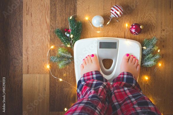 Fototapeta Top view of female feet in winter pajamas on digital scales or weight scale on wooden background surrounded with Christmas lights and decoration. Weight gain during holidays concept, vintage toned