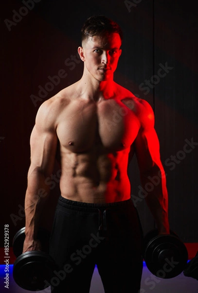 Fototapeta Muscular bodybuilder training biceps with dumbbells in red lighting.