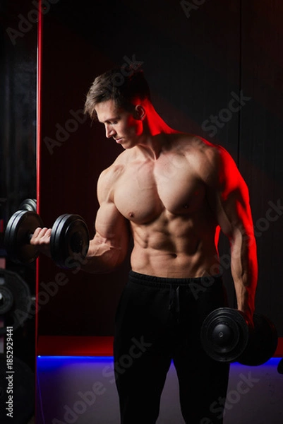 Obraz Muscular bodybuilder training biceps with dumbbells in red lighting.