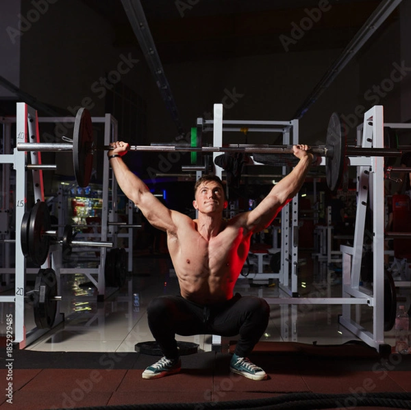 Fototapeta Muscular athlete performing wide-grip overhead barbell squat in a gym.