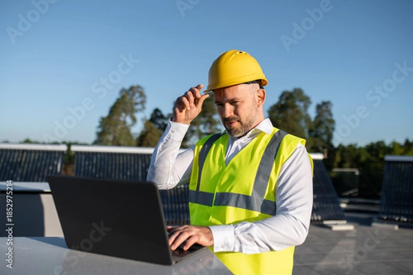 Obraz Engineer in helmet working on laptop near solar panels outdoors