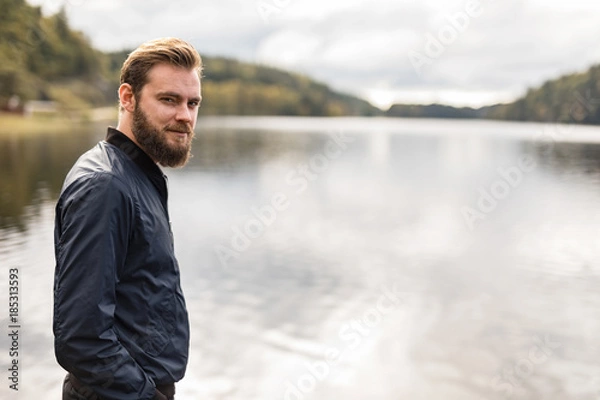 Fototapeta Laid back man with beard standing against a big lake looking at camera outdoors on a sunny day.