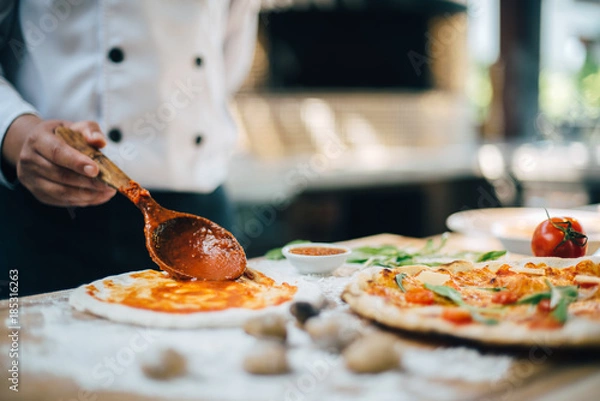 Fototapeta Fresh Italian pizza making process with hand of chef put tomato sauce on dough and pizza oven in background