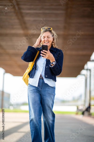 Obraz Smiling mature woman using smartphone while walking outside