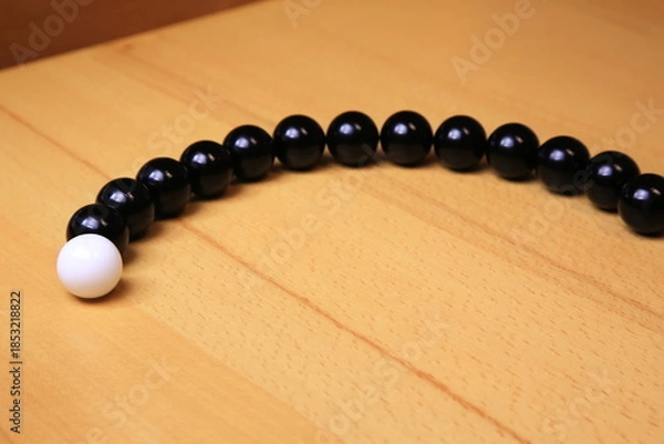 Fototapeta Eine weiße Kugel steht getrennt vor einer geschwungenen Reihe schwarzer Kugeln auf Holz; der starke Hell-Dunkel-Kontrast und die reduzierte Komposition betonen Individualität.