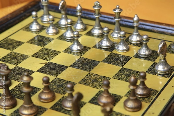 Fototapeta Metallisches Schachspiel auf gelb-schwarzem Brett, stilvoll arrangiert auf einem Holztisch. Silberne und bronzene Figuren in Startposition – ein Hauch von Eleganz und Strategie.