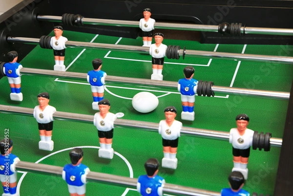 Fototapeta Kicker mit Überraschung: Ein Ei liegt zentral auf dem grünen Spielfeld, flankiert von blauen und weißen Kunststoffspielern auf Metallstangen; ungewöhnlicher, stiller Moment im Tischfußball.