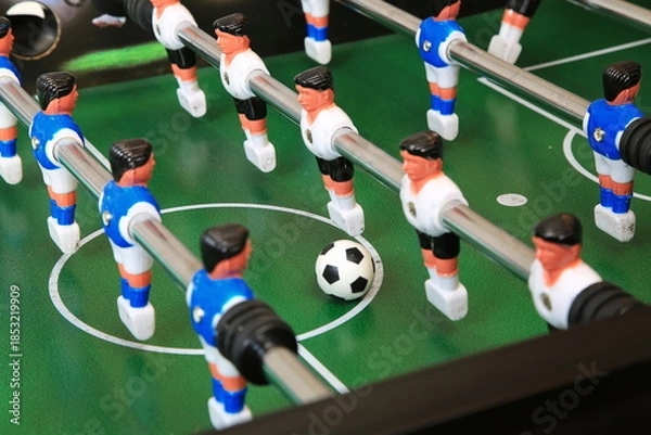 Fototapeta Kicker-Nahaufnahme: Ball liegt zentral auf dem grünen Spielfeld, flankiert von blauen und weißen Kunststoffspielern auf Metallstangen; ein dynamischer Moment im Tischfußballspiel.